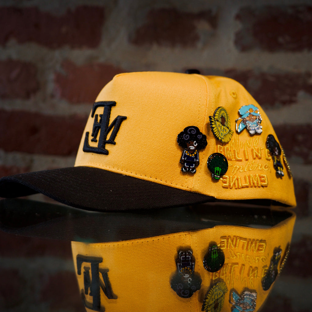 Emline yellow cap with black brim and various SPY BOY and space broccoli pins against a brick wall background