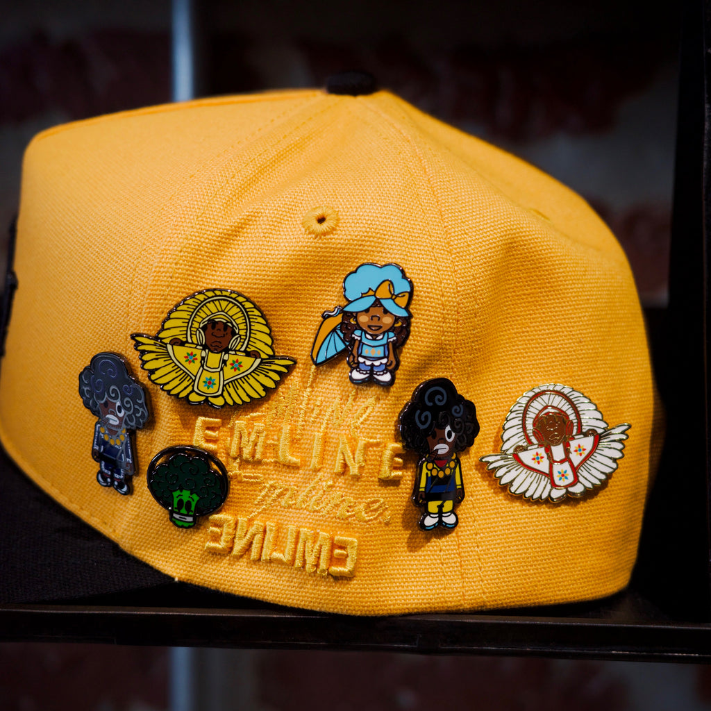 Emline yellow cap with black brim and various SPY BOY and space broccoli pins against a brick wall background