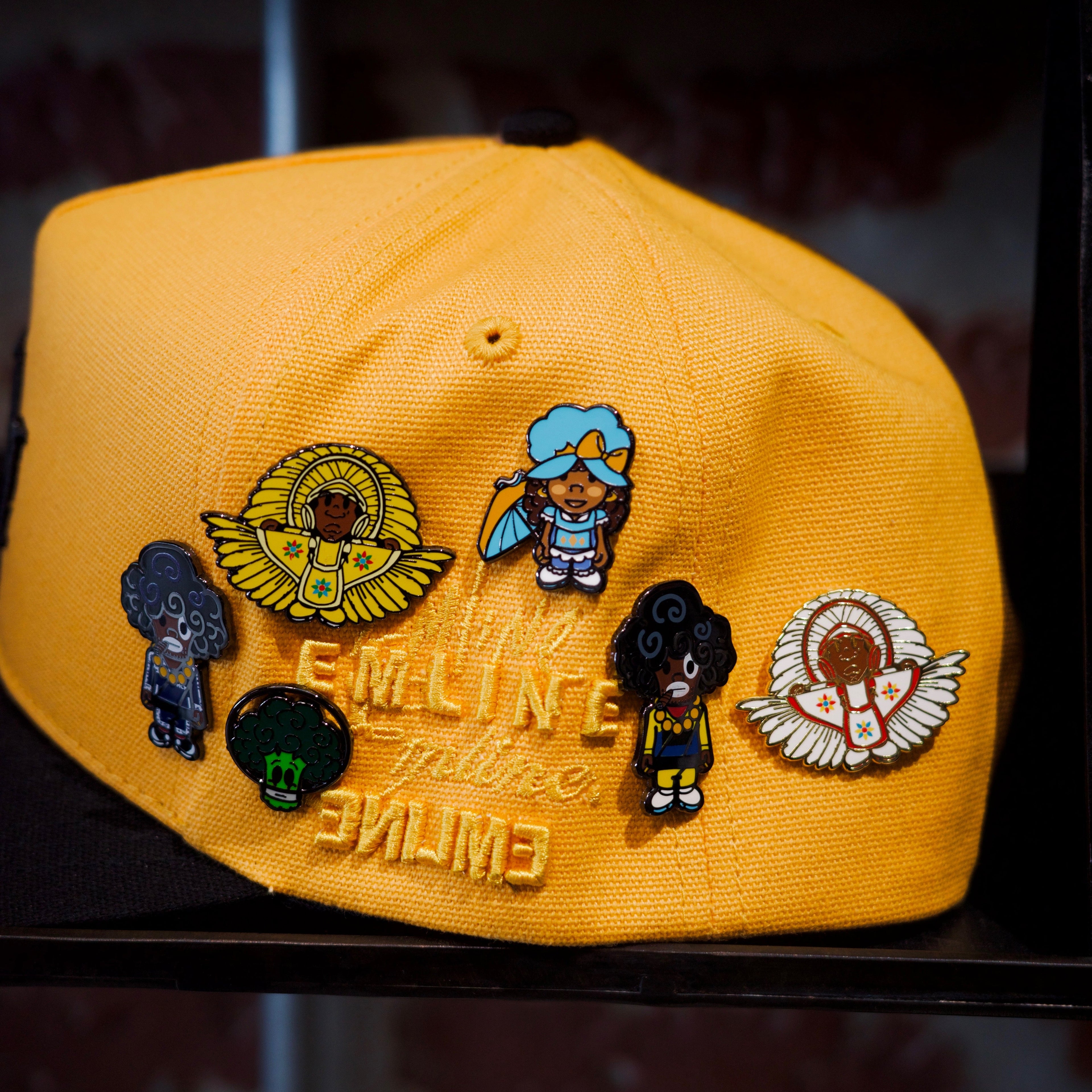 Emline yellow cap with black brim and various SPY BOY and space broccoli pins against a brick wall background